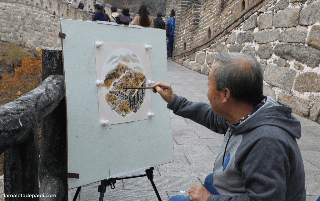 Un artista mayor pinta una escena del Gran Muralla China en un caballete, mientras que personas caminan de fondo.