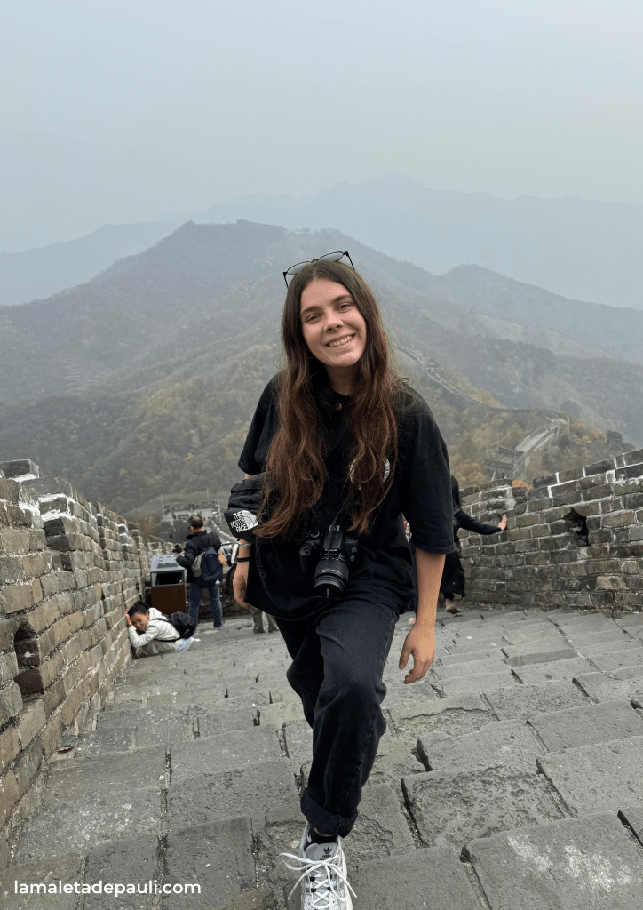 Una persona sonriente subiendo por la Gran Muralla China, acercándose a la cámara con un fondo de montañas y niebla.