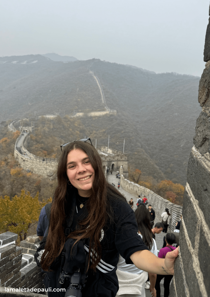 Una joven sonriente con cabello largo y oscuro posa frente a la Gran Muralla China, con un paisaje montañoso y neblinoso de fondo.