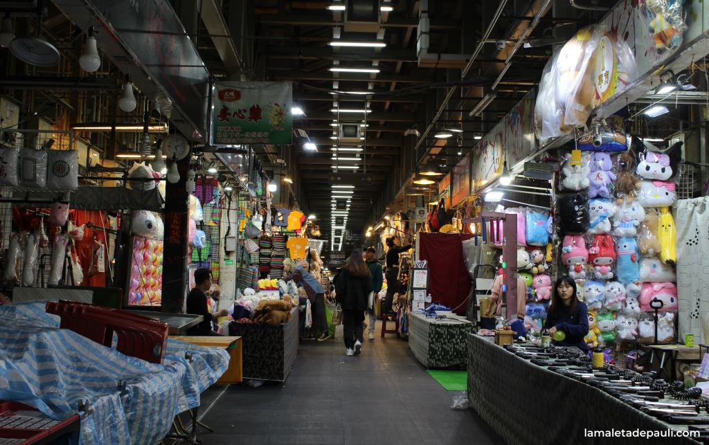 Un mercado nocturno con múltiples puestos que ofrecen peluches y artículos variados, iluminado con luces brillantes.