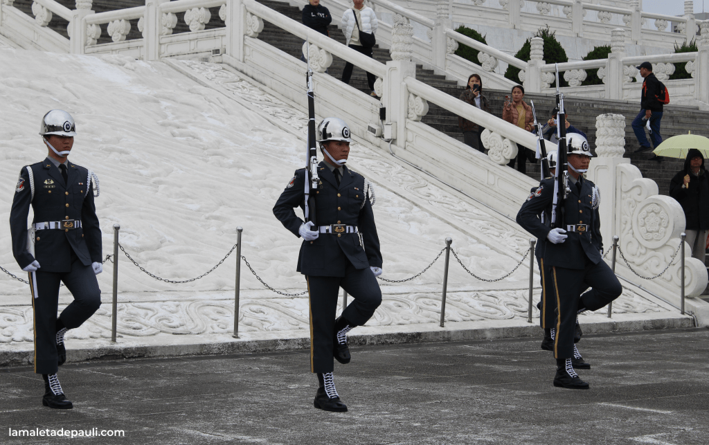 Tres guardias de honor en uniforme militar, realizando un desfile ceremonial, con un fondo de escaleras y personas observando.