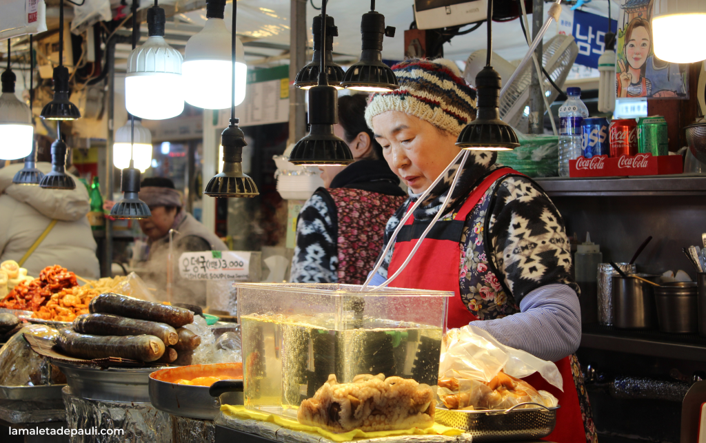 Una mujer de mediana edad en un mercado callejero, vistiendo un suéter de colores y un gorro de lana, está preparando alimentos. En su puesto hay una variedad de mariscos y otros platillos, con luces colgantes iluminando la escena.