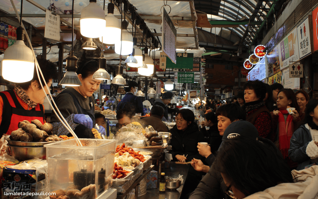 Mercado de comida con vendedores y clientes, donde se ofrecen mariscos y otros platillos. Hay luces colgantes y mucha actividad alrededor.