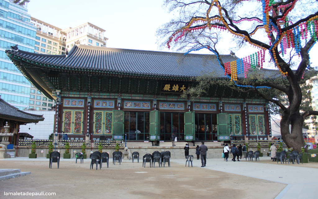 Templo tradicional asiático con arquitectura detallada, rodeado de un entorno urbano moderno. Personas caminando y sentadas en sillas frente al templo, con un árbol decorado con luces coloridas al lado.