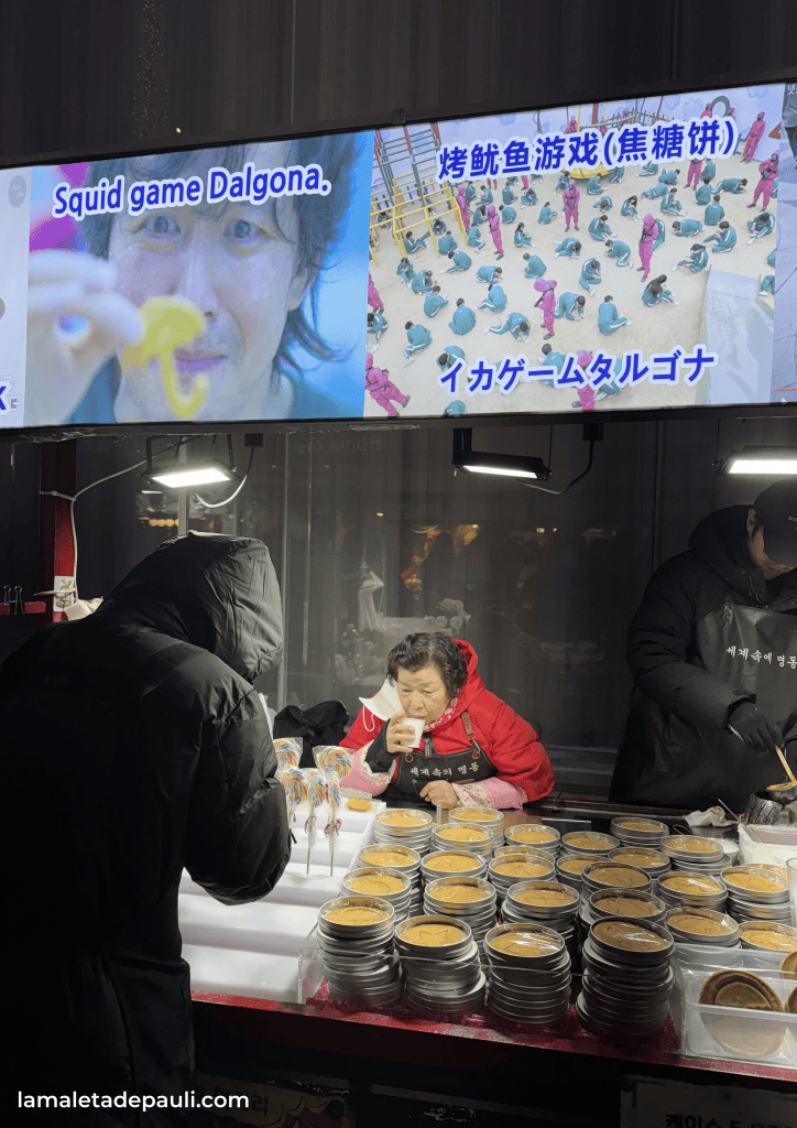 Una mujer sentada en un puesto de comida, disfrutando de una bebida, mientras hay bandejas de Dalgona y caramelos de colores a su alrededor. En la parte superior, hay una pantalla que muestra imágenes relacionadas con el juego del calamar (Squid Game).