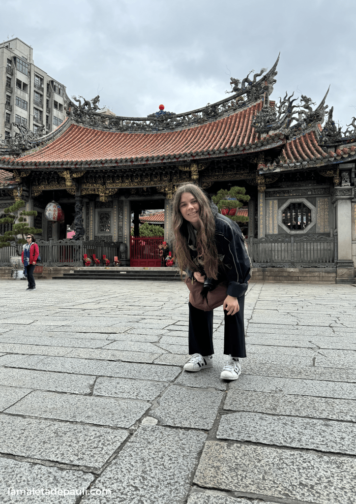 Una mujer sonriente se agacha frente a un templo tradicional en una plaza de piedra, con edificios modernos al fondo y un cielo nublado.