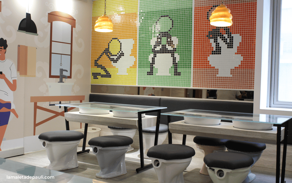 Interior de un restaurante con mesas de vidrio y asientos en forma de inodoro, decorado con mosaicos coloridos en la pared que representan figuras sentadas en inodoros y elementos de baño.
